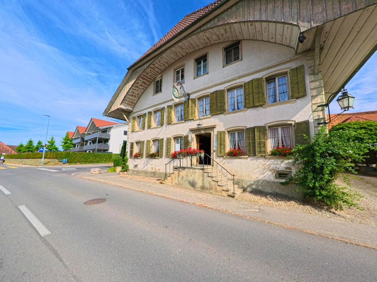 In Murten (3215, Kanton Freiburg) steht an einer leicht befahrenen Strasse ein traditionelles Gasthaus mit grünen Fensterläden, Blumenkästen und geschwungenem Vordach.