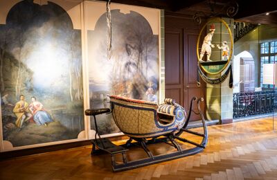 Im Musee Historique de Vevey (1800, Kanton Waadt) steht ein reich verzierter antiker Schlitten vor Wandgemaelden mit historischen Szenen und einem alten Wirtshausschild.