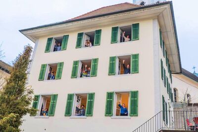 Im Museum Grenchen (2540, Kanton Solothurn) winken mehrere Personen fröhlich aus den Fenstern eines hellen Gebäudes mit grünen Fensterläden.