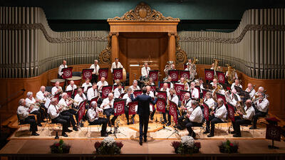 Das Musik-Corps Alte Garde Winterthur gibt ein Konzert auf einer Bühne vor einer grossen Konzertorgel. Unter der Leitung des Dirigenten spielt das Blasorchester ein vielseitiges Repertoire und pflegt die traditionelle Blasmusik.
