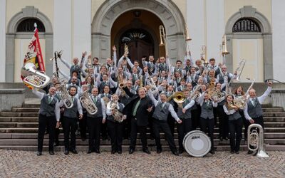 Vor der Kirche in Andwil (9402, Kanton St.Gallen) jubelt die Musikgesellschaft Andwil-Arnegg mit erhobenen Blasinstrumenten auf der Treppe für ein Gruppenfoto.
