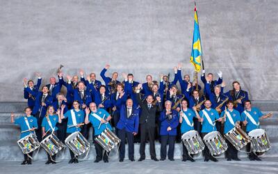 Die Mitglieder der Musikgesellschaft Maienfeld aus Maienfeld (7304, Kanton Graubünden) posieren gemeinsam mit Blasinstrumenten, Trommeln und Vereinsfahne für ein Gruppenfoto.