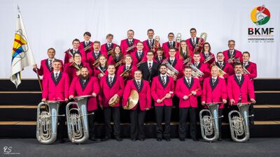 Die Musikgesellschaft Richenthal-Langnau aus Richenthal (6263, Kanton Luzern) posiert in roten Anzügen mit Instrumenten und einer Flagge für ein Gruppenfoto.