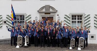 In Sins (5643, Kanton Aargau) steht eine Musikgesellschaft in Uniform mit Instrumenten vor einem Gebäude und posiert für ein Gruppenfoto.