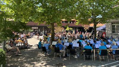 In Uetikon am See (8707, Kanton Zürich) spielt der Musikverein im Schatten grosser Bäume vor zahlreichem Publikum auf einem sonnigen Platz.