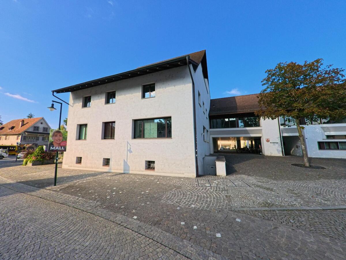 Auf einem weitläufigen Platz in Muttenz (4132, Kanton Basel-Landschaft) steht ein modernes Gemeindegebäude neben einem schlichten Wohnhaus, umgeben von Kopfsteinpflaster.