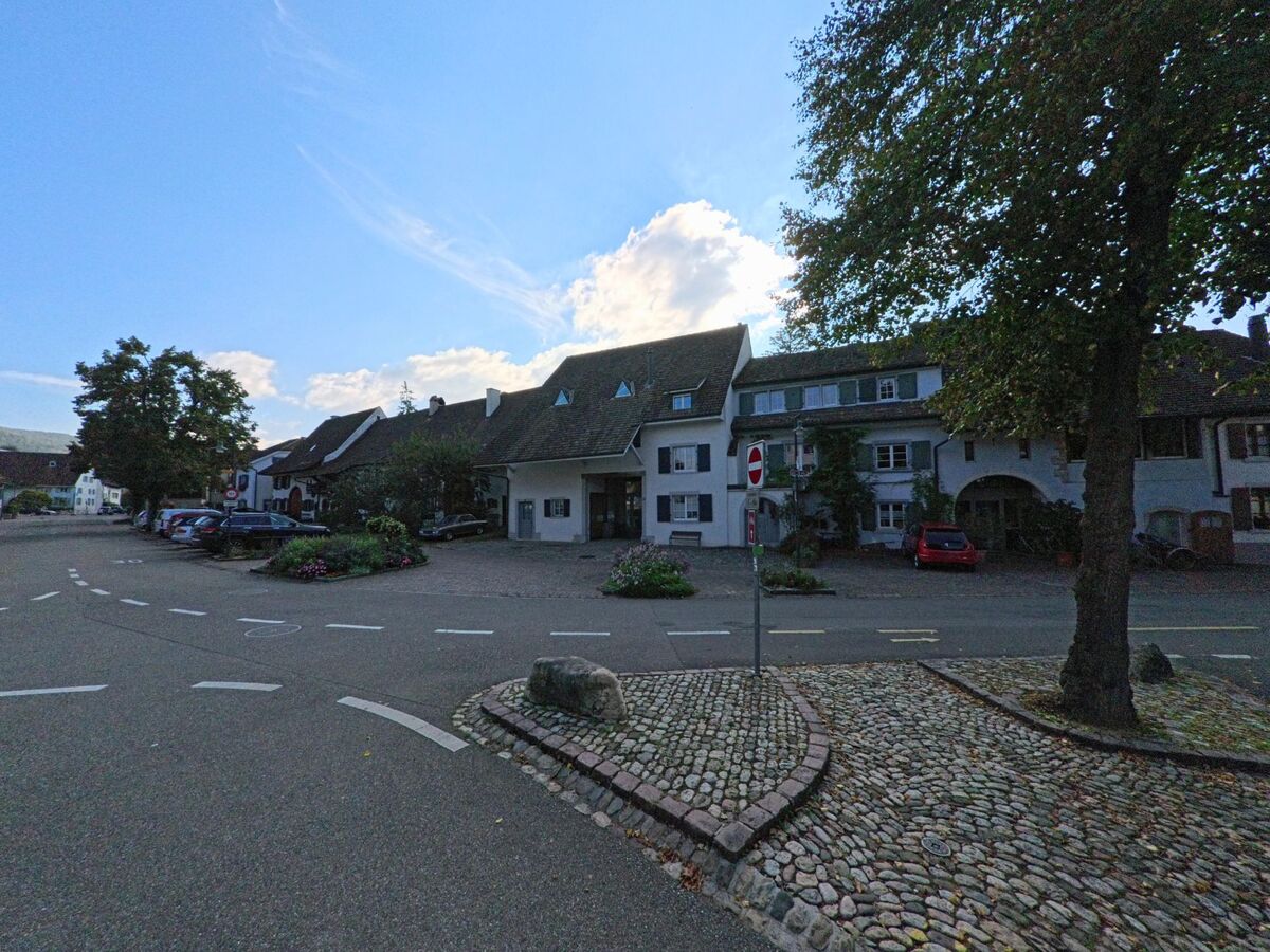 In Muttenz (4132, Kanton Basel-Landschaft) säumen traditionelle Häuser mit begrünten Fassaden einen Platz, an dessen Rand eine Verkehrsinsel aus Kopfsteinpflaster liegt.