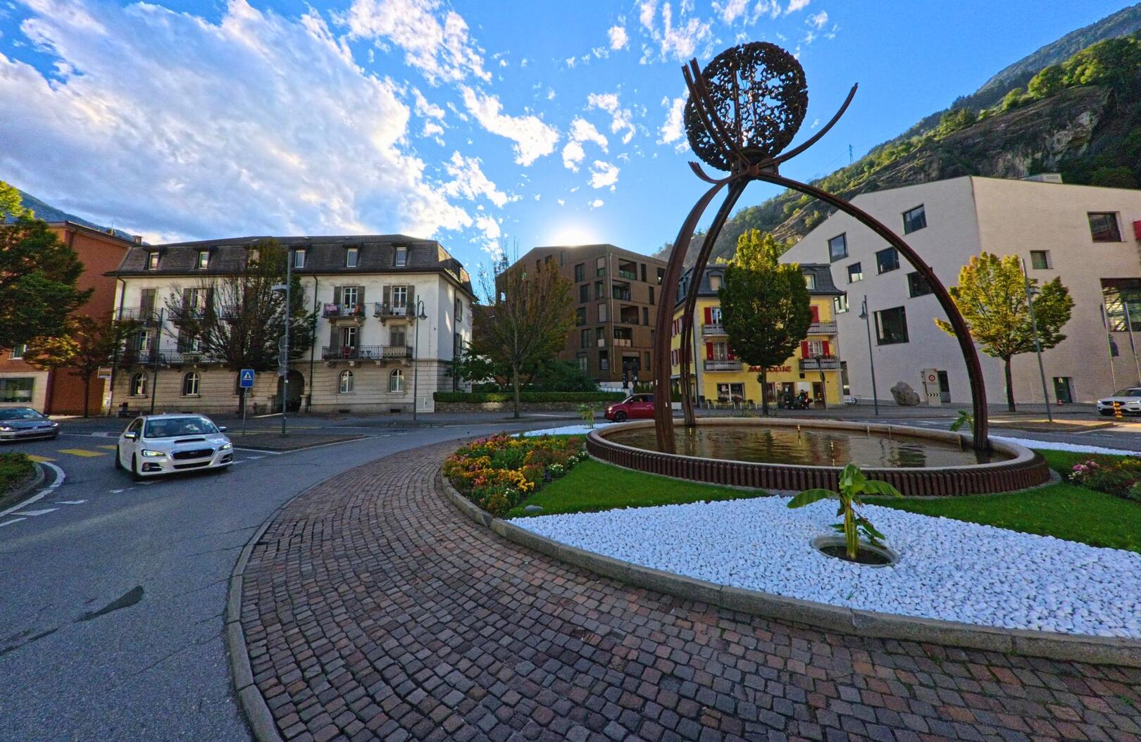Ein moderner Brunnen mit kunstvoller Metallskulptur steht in einem Kreisverkehr von Naters (3900, Kanton Wallis), umgeben von Blumenbeeten und historischen Gebaeuden.