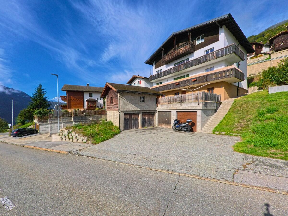 Am Hang von Naters (3903, Kanton Wallis) stehen mehrere Wohnhaeuser mit Balkonen und Garagen, waehrend im Hintergrund die Berge sichtbar sind.