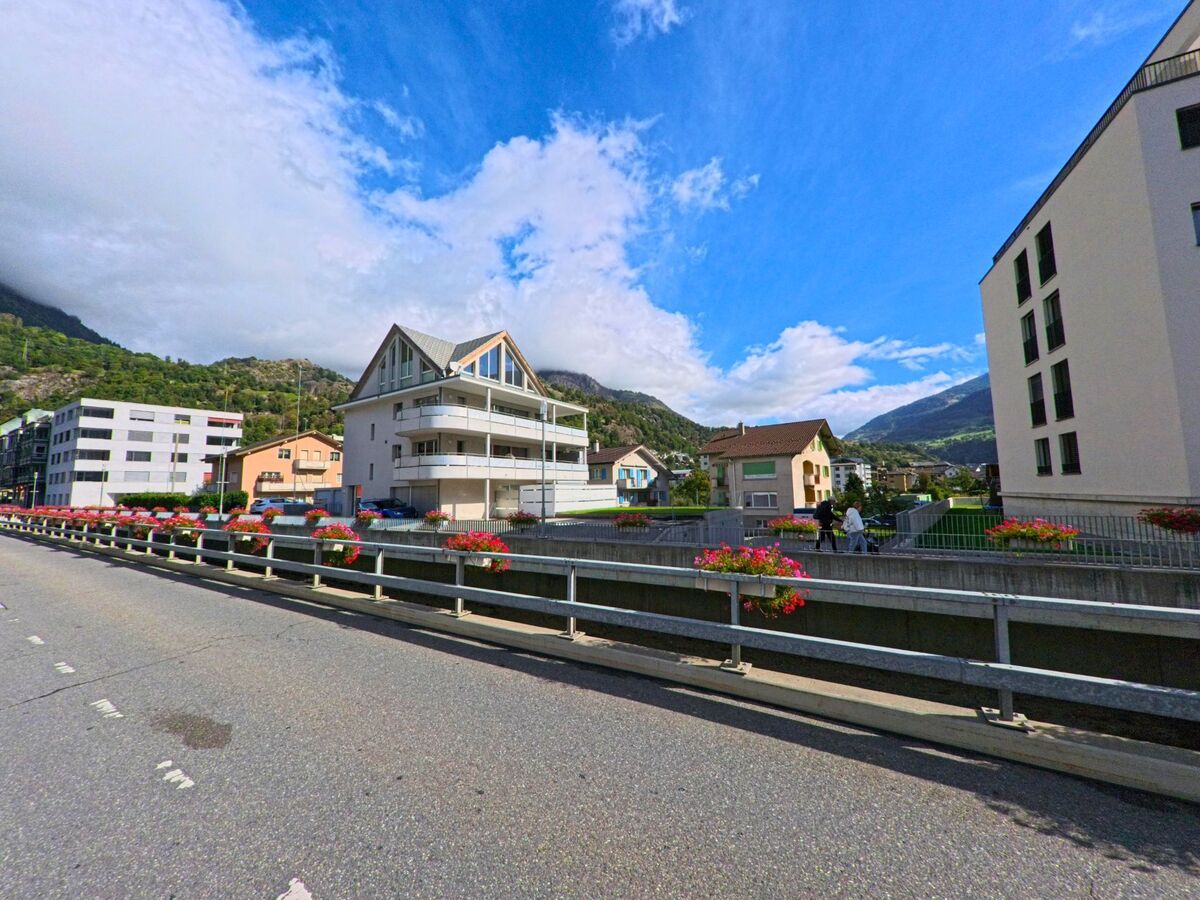 Von einer mit Blumenkisten geschmueckten Bruecke in Naters (3904, Kanton Wallis) blickt man auf moderne und traditionelle Wohnhaeuser vor bewaldeten Huegeln.