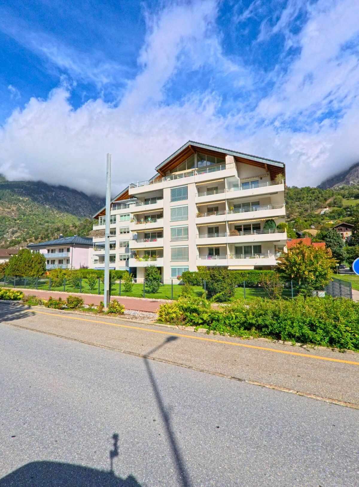 Ein mehrgeschossiges Wohnhaus mit grossen Balkonen und Satteldach steht in Naters (3904, Kanton Wallis) vor einer Kulisse aus bewaldeten Bergen und Wolken.