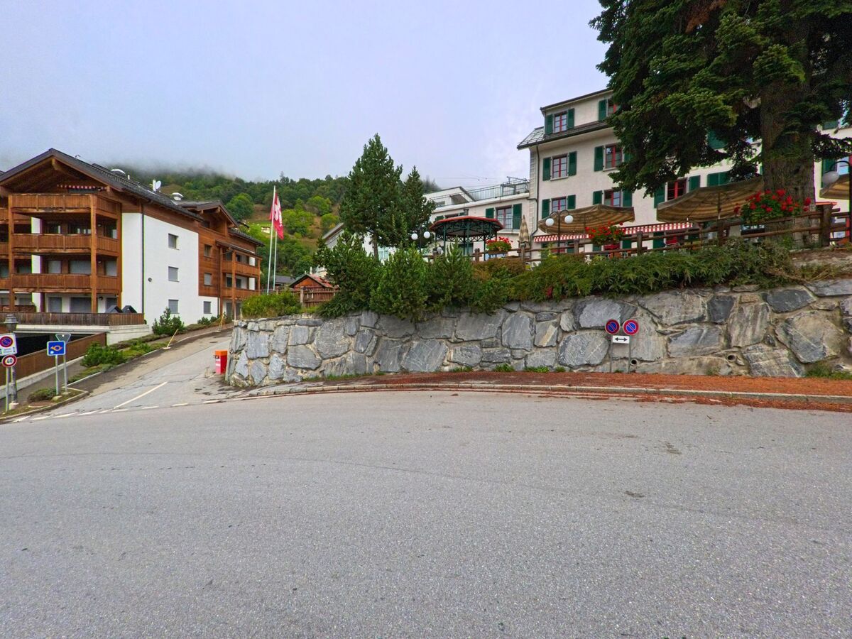 Vor einer steinernen Stuetzmauer in Naters (3914, Kanton Wallis) stehen Wohnhaeuser und ein Hotel mit roten Blumenkasten, daneben wehen zwei Schweizer Fahnen.