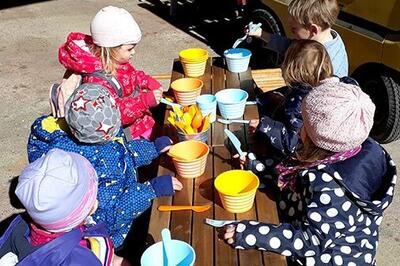 In Murten (3285, Kanton Freiburg) sitzen mehrere Kinder an einem Holztisch im Freien und spielen gemeinsam mit bunten Schalen und Werkzeugen.