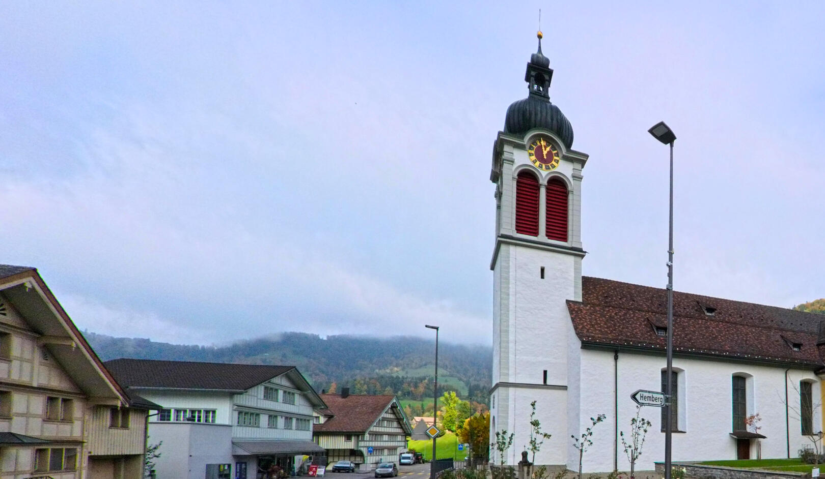 Im Zentrum von Neckertal (9122, Kanton St. Gallen) steht die weisse Kirche mit ihrem markanten Turm, waehrend im Hintergrund bewaldete Huegel unter leichtem Nebel liegen.