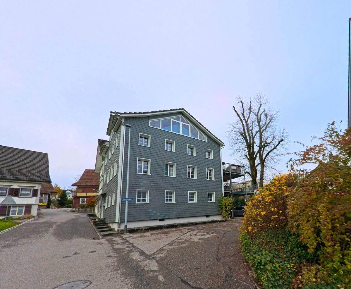 Ein Schindelhaus mit heller Fassade steht in einer kleinen Platzsituation von Neckertal (9122, Kanton St. Gallen).
