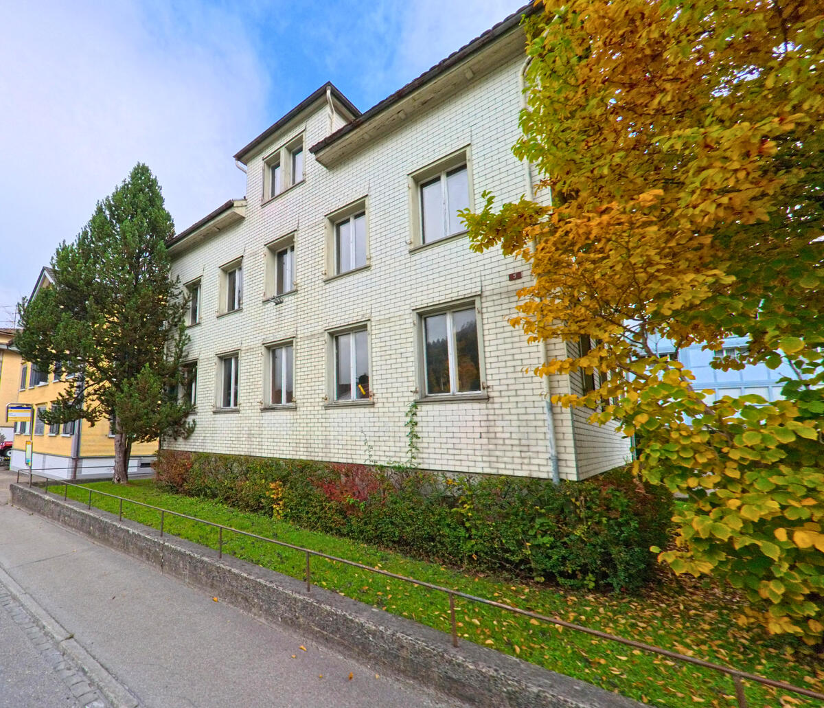 Ein weiss verputztes Gebaeude mit klinkerartiger Fassade liegt entlang einer Nebenstrasse in Neckertal (9122, Kanton St. Gallen).