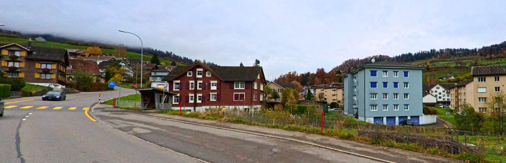 In Nesslau (9650, Kanton St. Gallen) verlaeuft eine Strasse an Wohnhaeusern und einem kleinen Unterstand vorbei, waehrend die Huegellandschaft im Hintergrund von tiefen Wolken bedeckt ist.