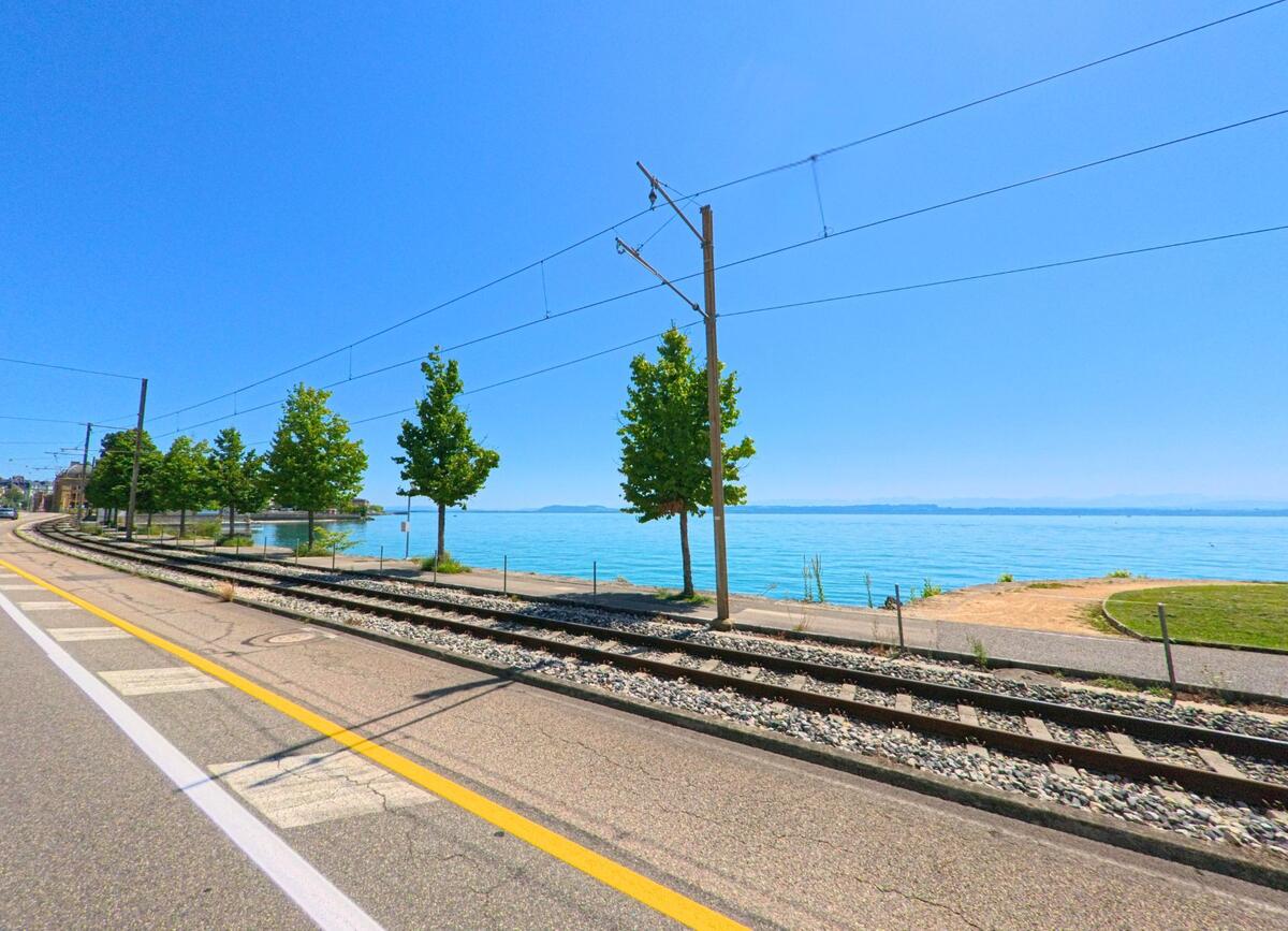 Entlang des Ufers in Neuchatel (2002, Kanton Neuenburg) verlaeuft eine Bahnstrecke parallel zur Strasse, dahinter liegt der See mit klarem Wasser unter blauem Himmel.