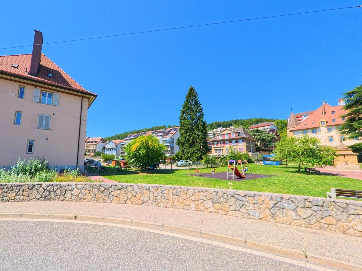 In Neuchatel (2034, Kanton Neuenburg) befindet sich eine kleine Parkanlage mit Spielplatz, umgeben von Wohnhaeusern und einem historischen Gebaeude mit Turm.