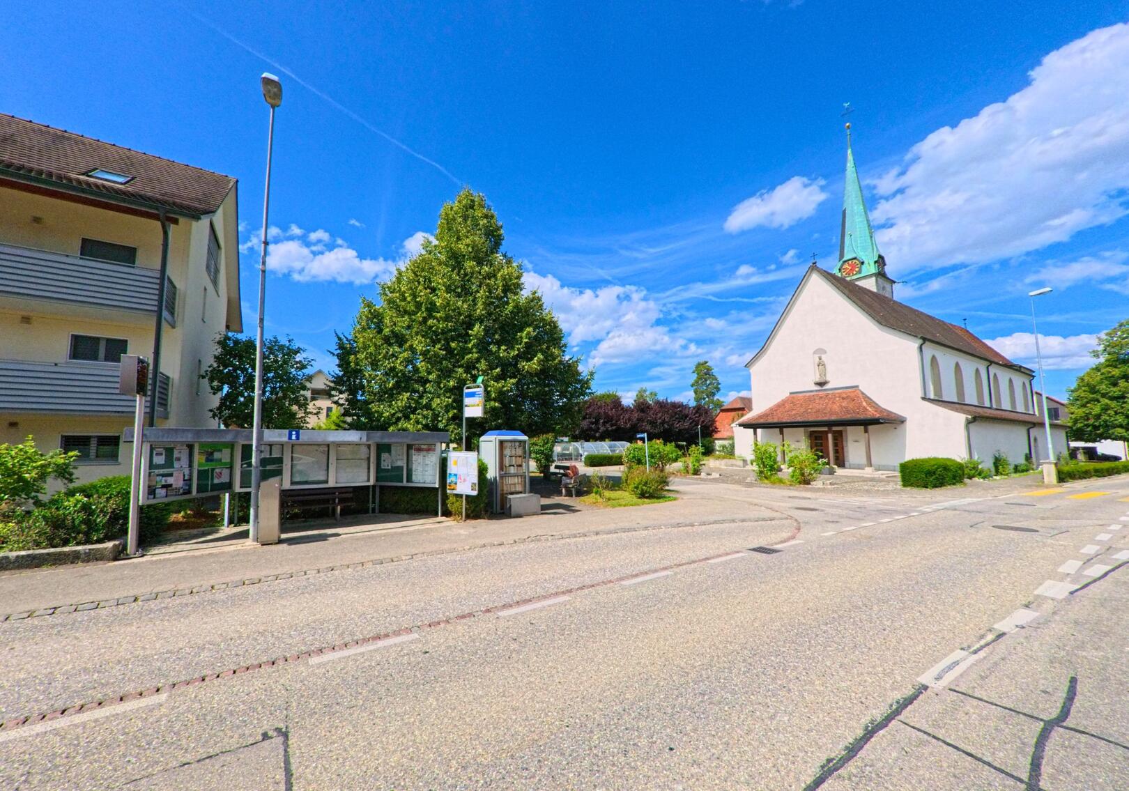 In Neuendorf (4623, Kanton Solothurn) befindet sich an einer Strassenecke ein kleiner Dorfplatz mit Bushaltestelle, öffentlichem Anschlagbrett und der Kirche mit grünem Turmdach.