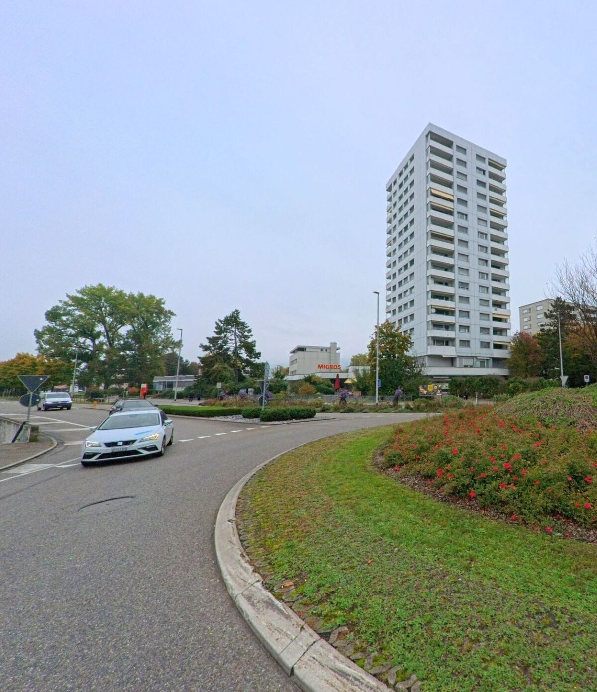 Ein hoher Wohnblock und ein Migros-Gebaeude praegen die Szenerie am Kreisverkehr in Neuenhof (5432, Kanton Aargau), umgeben von Gruenflaechen und Blumenbeeten.