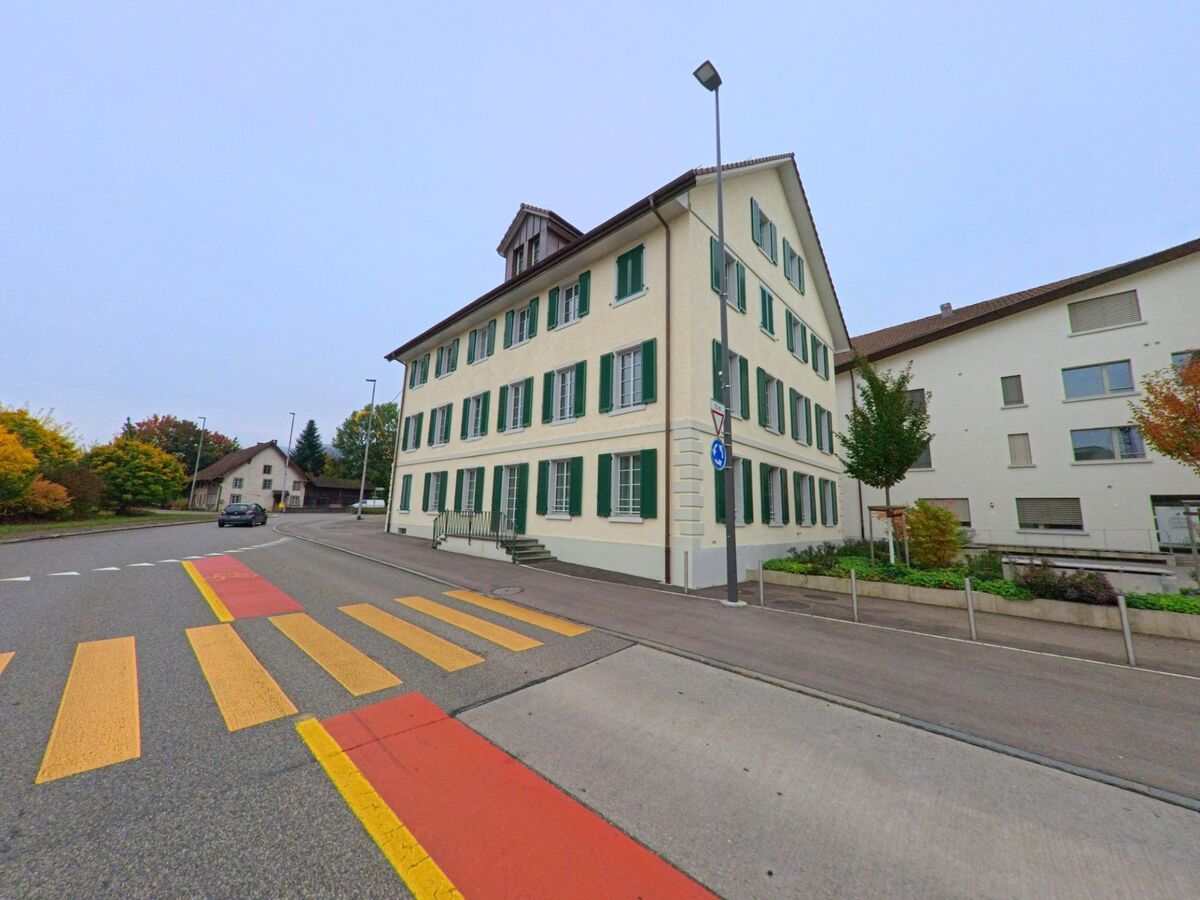 Ein helles Gebaeude mit gruenen Fensterlaeden steht an einer Strasse mit markiertem Fussgaengerstreifen in Neuenhof (5432, Kanton Aargau).