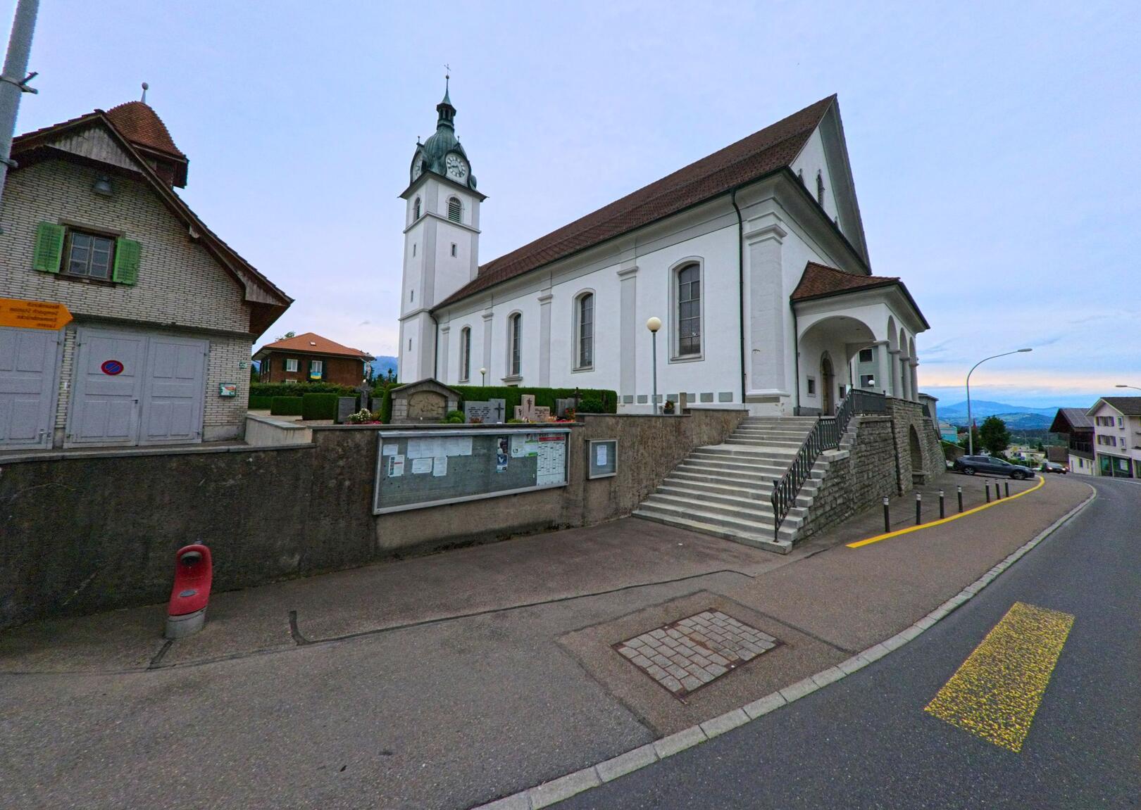 Die weisse Kirche von Neuenkirch (6016, Kanton Luzern) steht erhöht an der leicht abschüssigen Ortsdurchfahrt mit Blick auf das umliegende Hügelland.