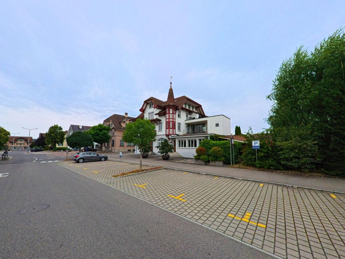 In Neuenkirch (6203, Kanton Luzern) steht ein markantes Gebäude mit Fachwerkfassade an einem ruhigen Platz mit Parkplätzen und Bäumen.