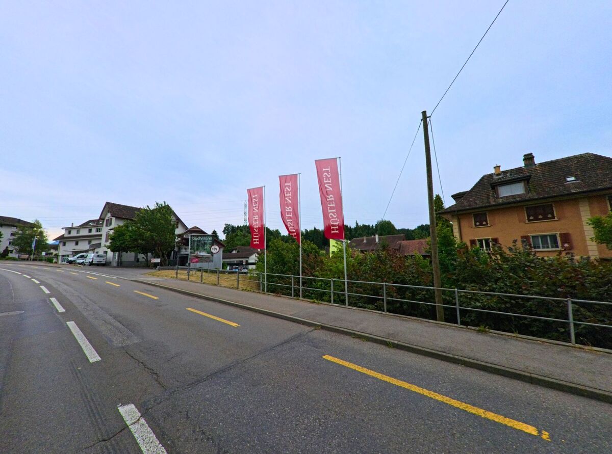 Entlang der Hauptstrasse in Neuenkirch (6206, Kanton Luzern) wehen rote Fahnen mit weissem Schriftzug vor einem Gebaeudekomplex mit Werbetafel und Parkflaeche.