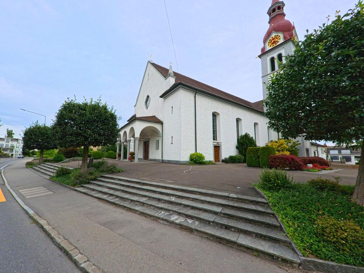 Die Kirche von Neuenkirch (6206, Kanton Luzern) mit rotem Turmhelm steht leicht erhoeht an einer bepflanzten Treppenanlage und ist von Bueschen und Baeumen eingerahmt.