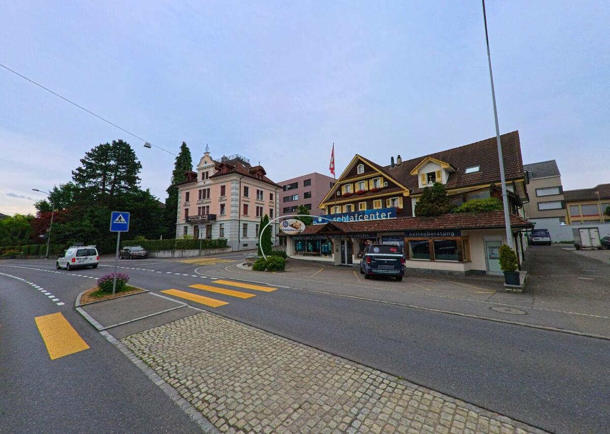 Direkt an einer Kreuzung in Neuenkirch (6206, Kanton Luzern) liegt ein Fachgeschaeft mit gelben Fensterlaeden neben einer historischen Villa und modernen Gebaeuden.