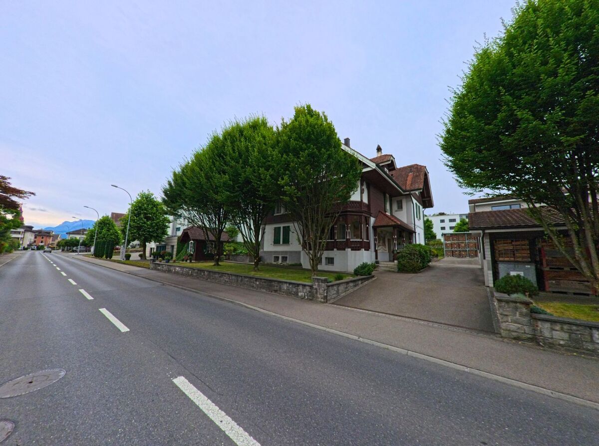 Entlang einer breiten Strasse in Neuenkirch (6206, Kanton Luzern) steht ein stattliches Wohnhaus mit Erker und Vorgarten, umgeben von einer Reihe hoher, dicht belaubter Baeume.
