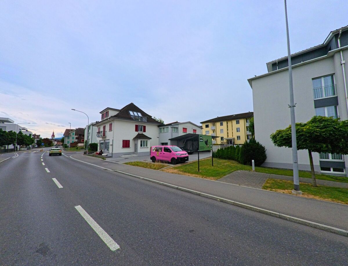 In Neuenkirch (6206, Kanton Luzern) saeumen Wohnhaeuser mit farbigen Fensterlaeden eine breite Strasse, auf der ein pinkfarbener Lieferwagen vor einem Schild parkiert ist.