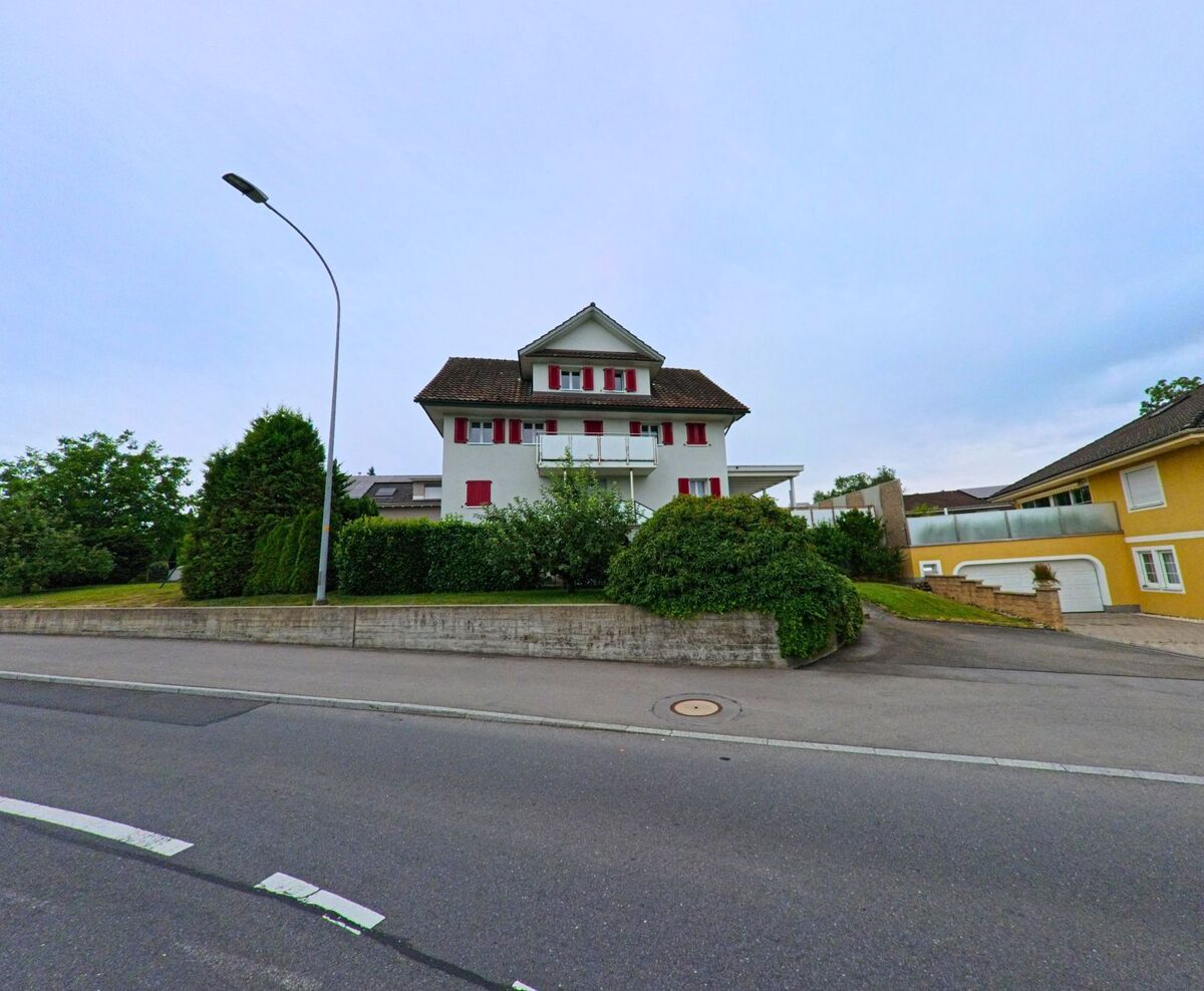 An einer ruhig gelegenen Strasse in Neuenkirch (6206, Kanton Luzern) steht ein weisses Wohnhaus mit roten Fensterlaeden, flankiert von gepflegtem Gruen und einem gelben Nachbarhaus.