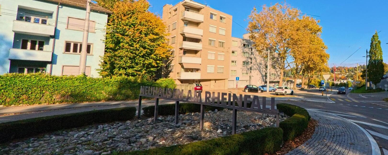 In Neuhausen am Rheinfall (8212, Kanton Schaffhausen) führt eine Rundstraße am begrünten Kreisverkehr entlang, mit modernen Wohngebäuden und einem klaren blauen Himmel im Hintergrund.