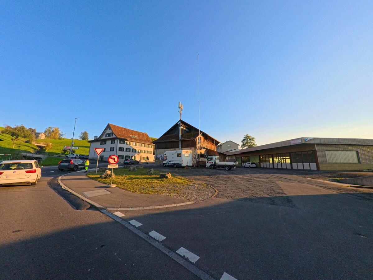 In Neuheim (6345, Kanton Zug) treffen ein traditionelles Wohnhaus, ein grosser Scheunenbau und ein Gewerbegebäude an einer belebten Strassenkreuzung aufeinander.