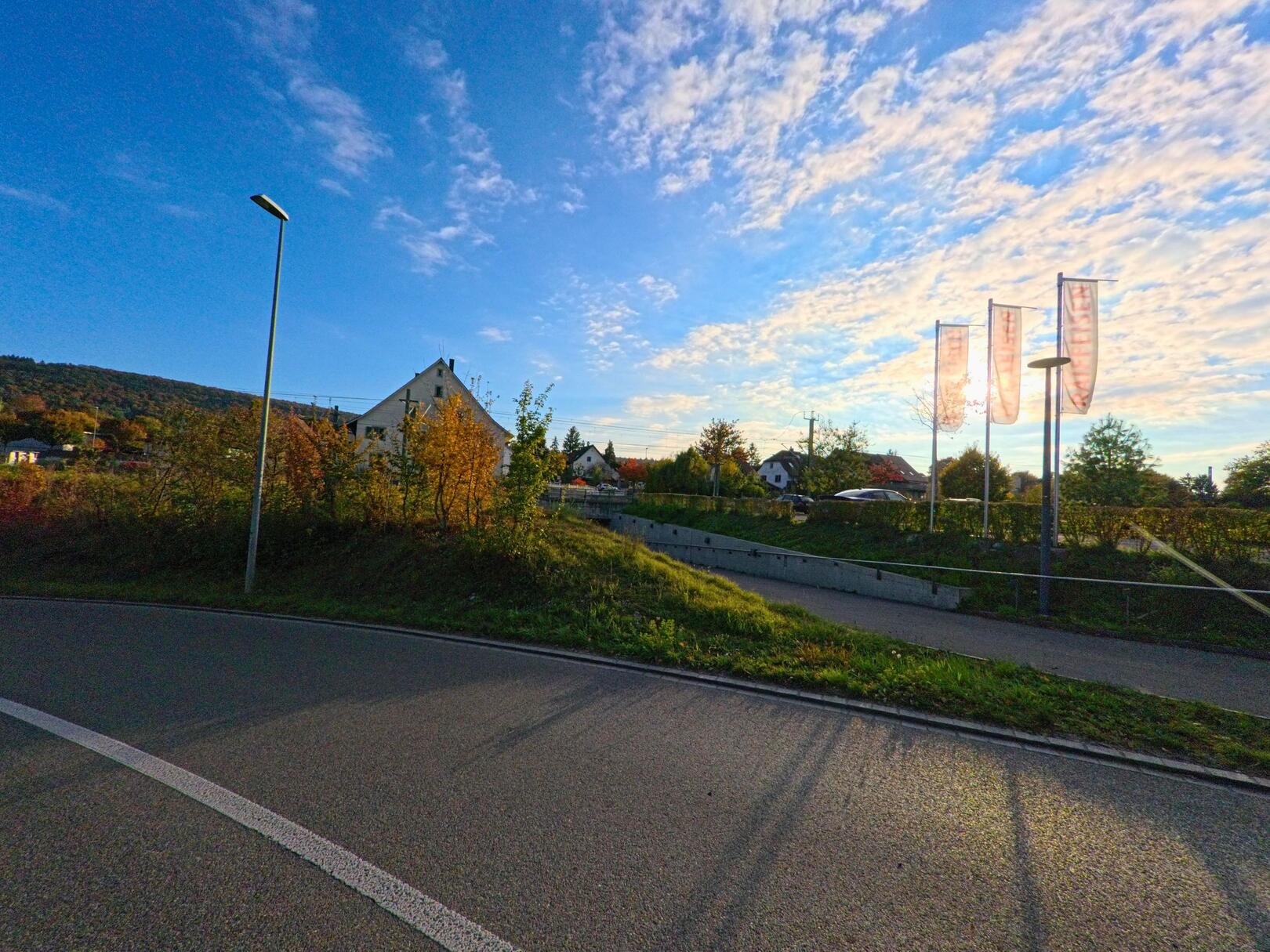 In Neunkirch (8213, Kanton Schaffhausen) zeigt das Bild eine malerische Szene mit herbstlich gefärbten Bäumen, einem kurvigen Weg und fahnengekrönten Masten vor einem Gebäude.