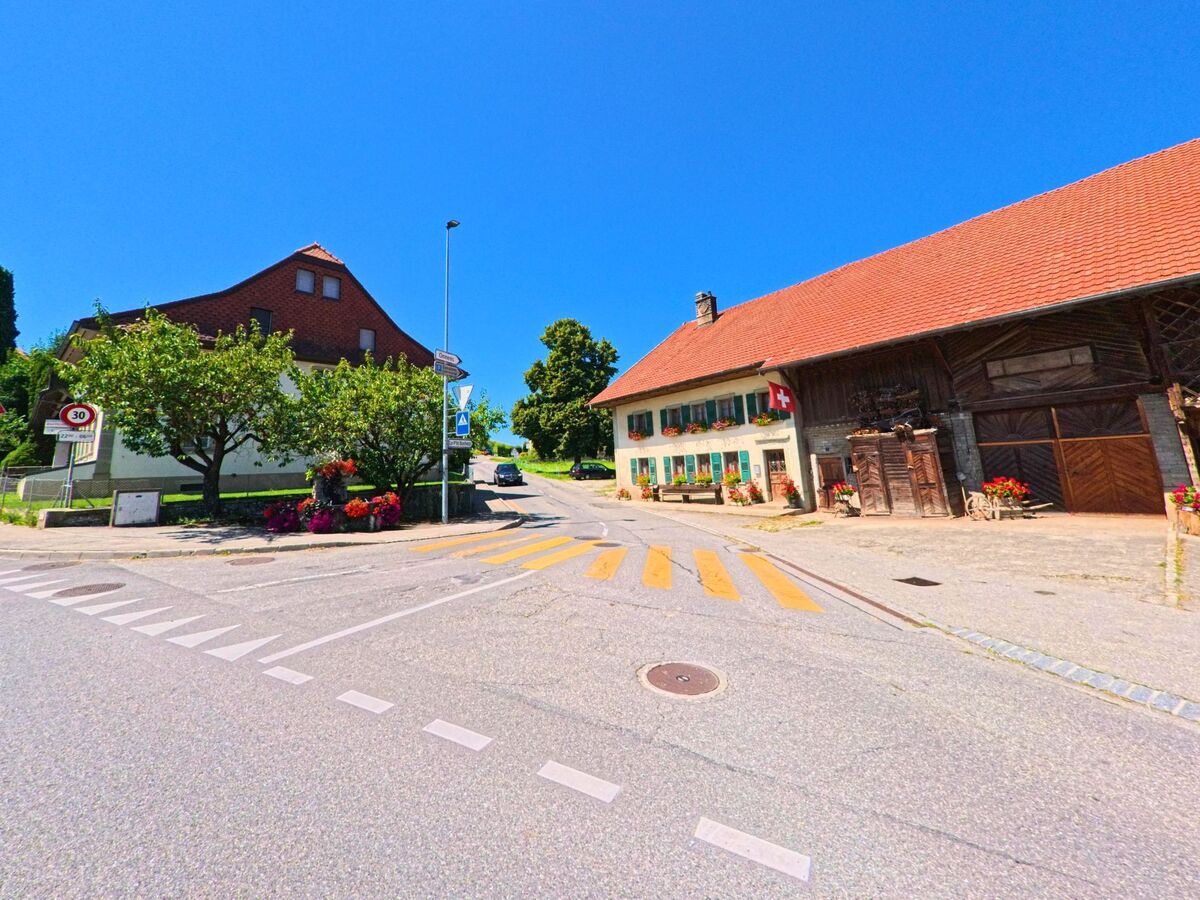 An einer Kreuzung in Neyruz (1740, Kanton Freiburg) steht ein traditionelles Bauernhaus mit Schweizer Flagge und blumengeschmückten Fenstern.