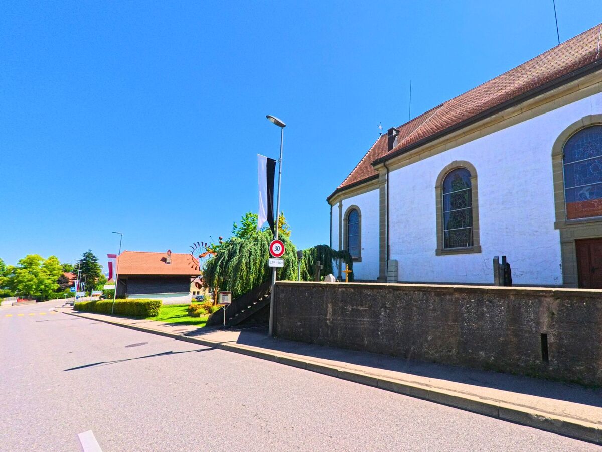 An einer Strasse in Neyruz (1740, Kanton Freiburg) steht eine Kirche mit weisser Fassade und grossen Rundbogenfenstern.