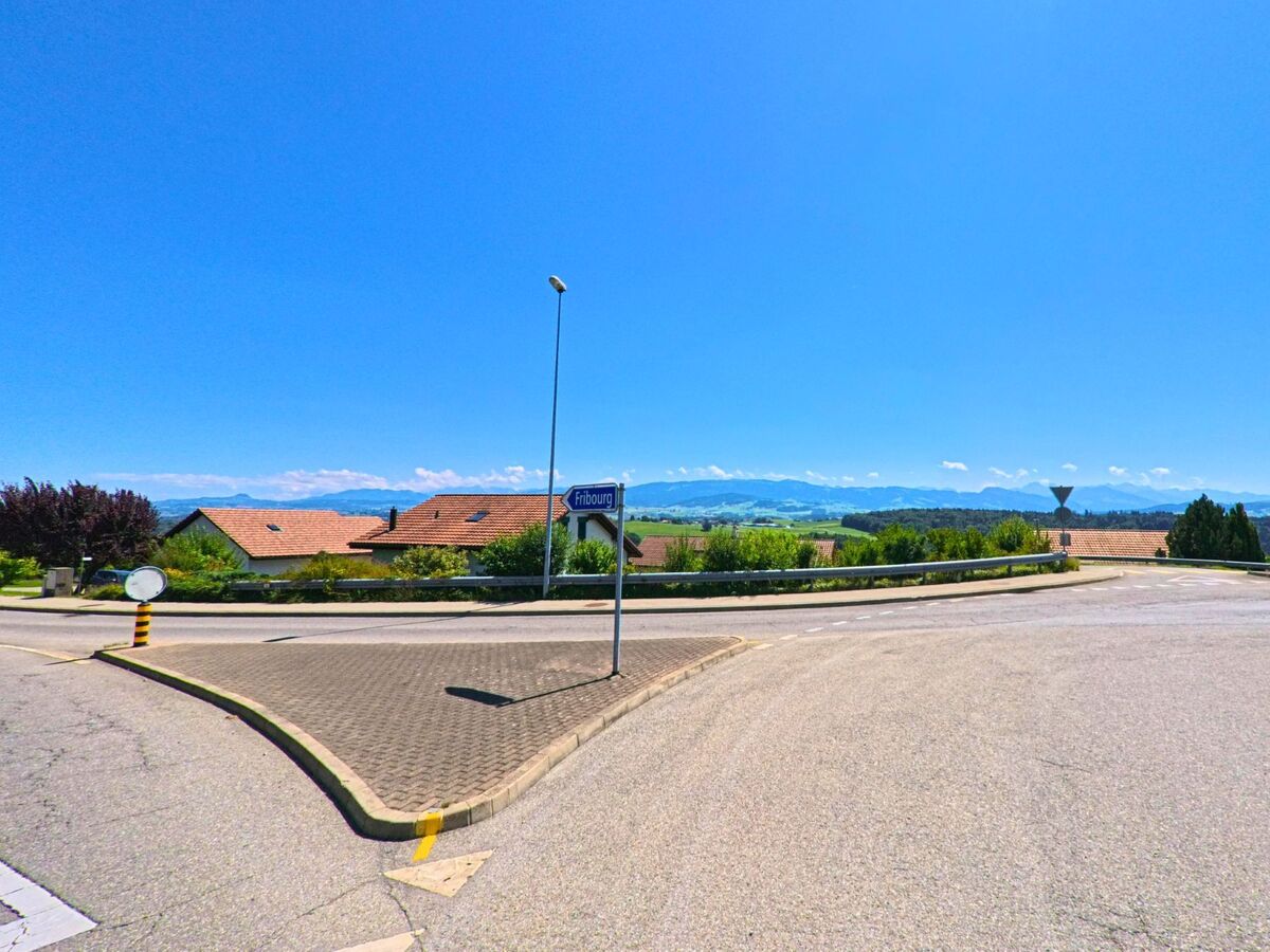 Von einer Strassenkreuzung in Neyruz (1740, Kanton Freiburg) öffnet sich der Blick über Dächer und Grünflächen bis zu den Alpen.