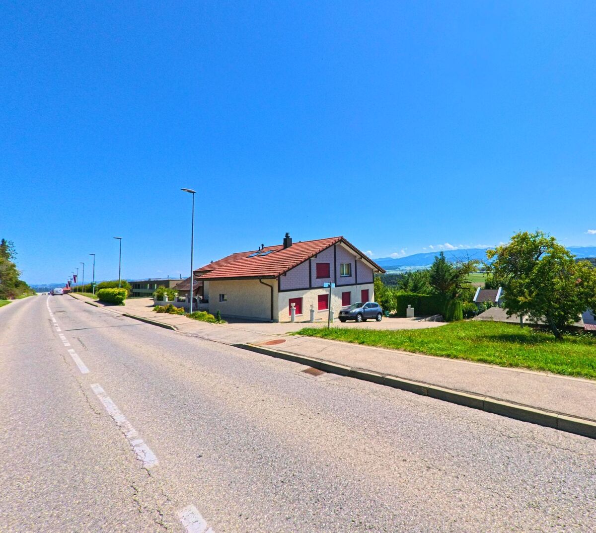 Entlang einer breiten Strasse in Neyruz (1740, Kanton Freiburg) stehen Einfamilienhäuser mit Blick auf die entfernten Alpen.