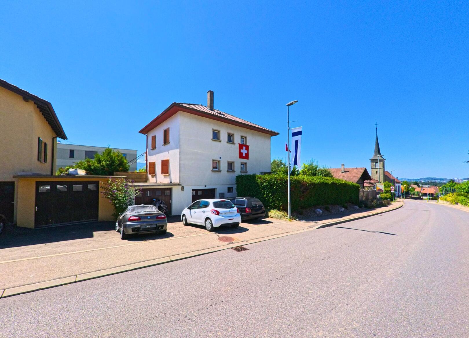 An einer Hauptstrasse in Neyruz (1740, Kanton Freiburg) steht ein Wohnhaus mit Schweizer Flagge, im Hintergrund ist der Kirchturm der Dorfkirche sichtbar.