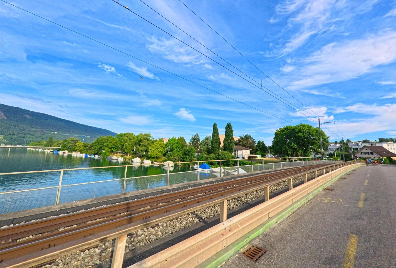 In Nidau (2560, Kanton Bern) führt die Strasse entlang der Eisenbahngleise, mit einem Blick auf das ruhige Gewässer und die umliegenden grünen Hügel.