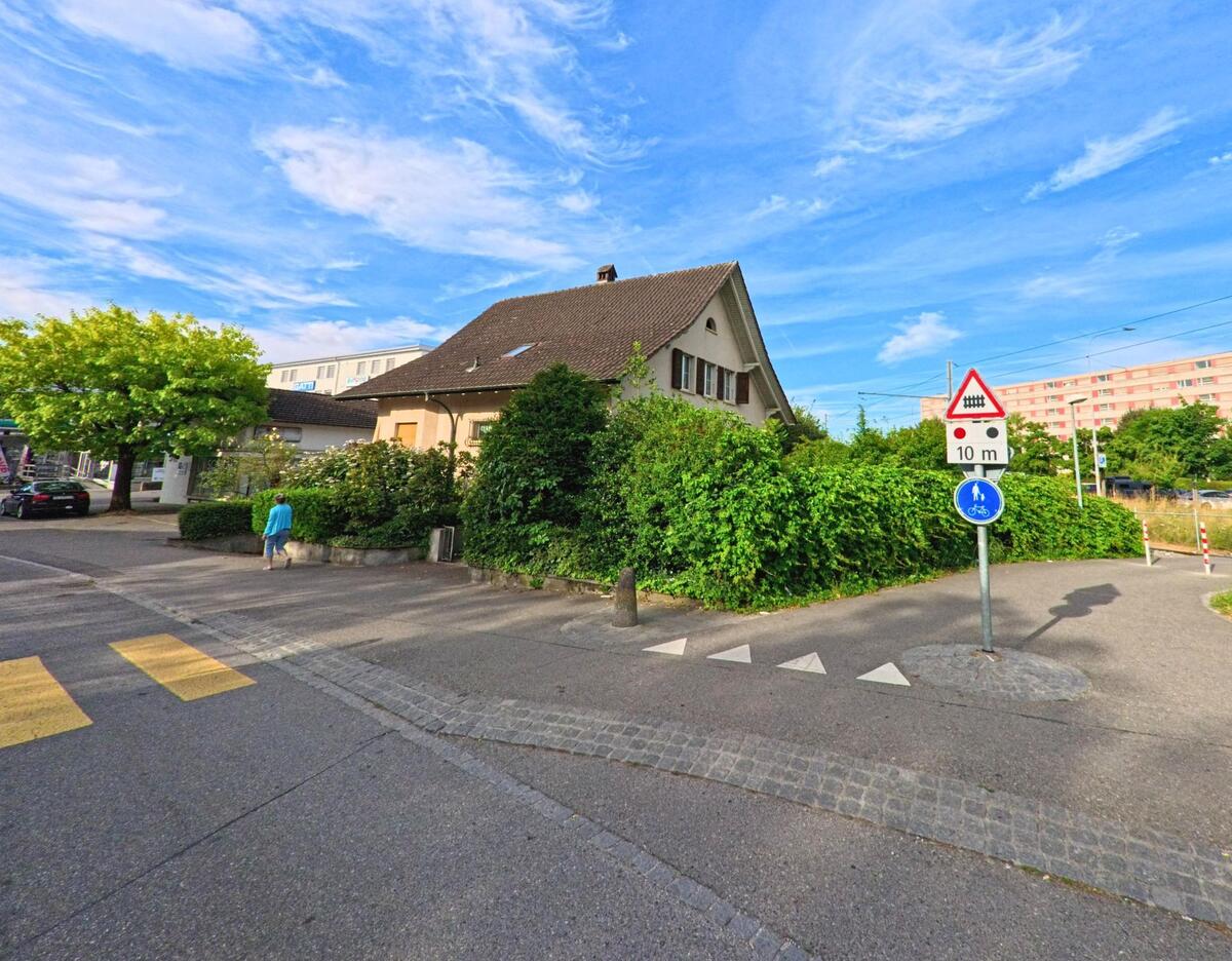 In Nidau (2560, Kanton Bern) führt die Straße an einem kleinen Haus und grünen Büschen vorbei, während Verkehrsschilder und Schranken den Weg zum nahegelegenen Bahnübergang kennzeichnen.