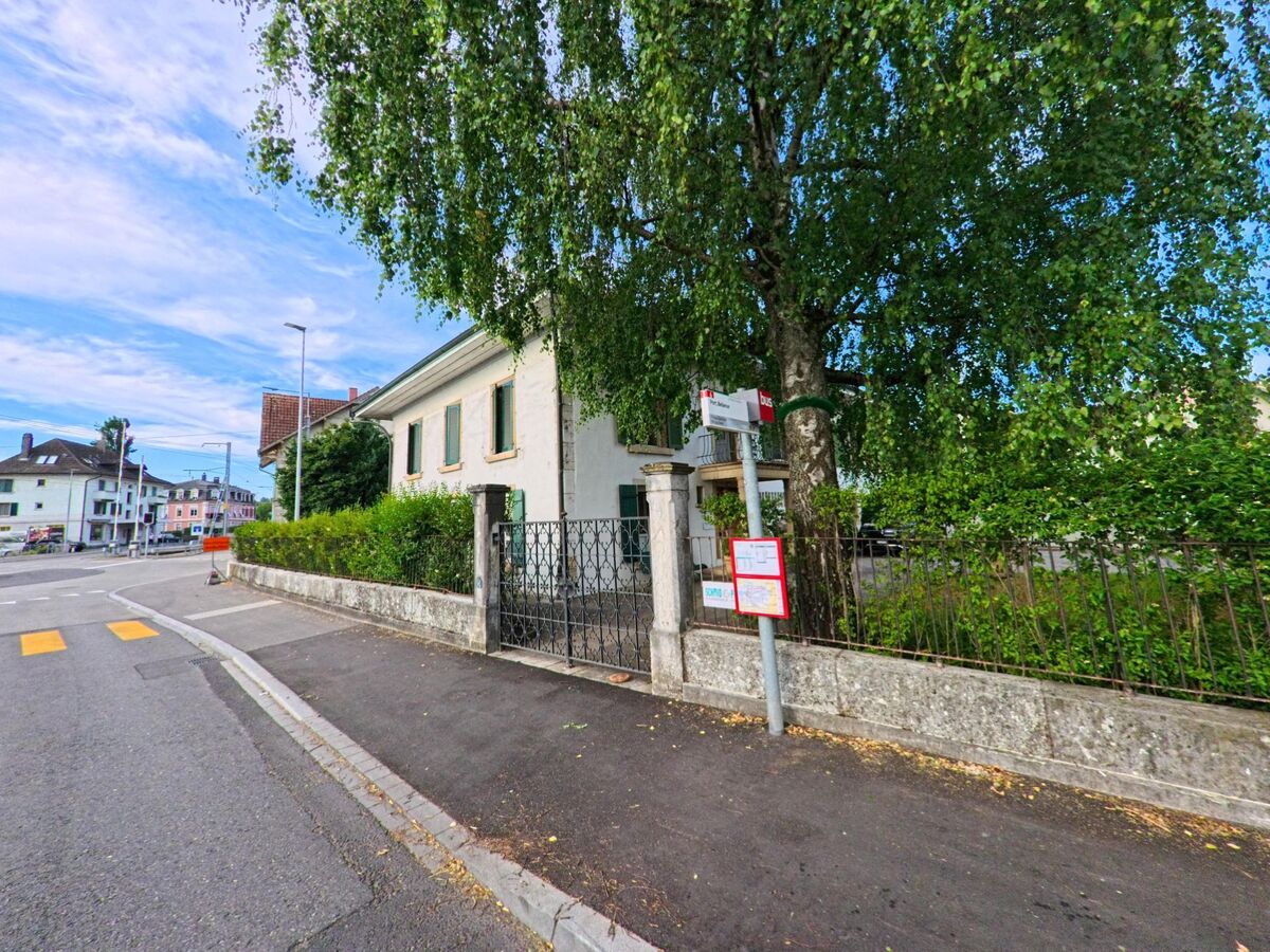 In Nidau (2560, Kanton Bern) ist eine ruhige Straßenszene mit einem historischen Gebäude und einer Bushaltestelle, die von grünen Bäumen und Büschen umrahmt wird.