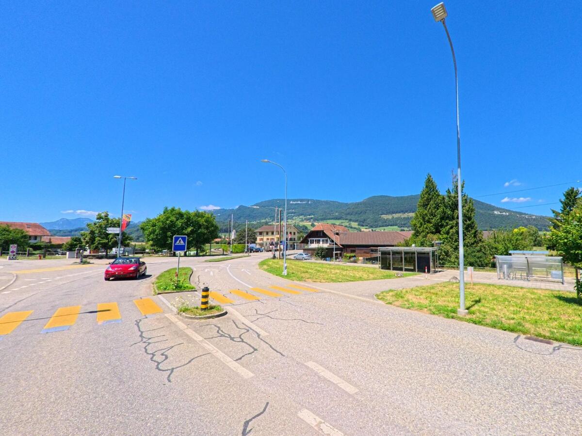 Eine breite Strasse mit Fussgaengerstreifen fuehrt in Niederbipp (4704, Kanton Bern) an einer Bushaltestelle vorbei in Richtung bewaldeter Huegel.