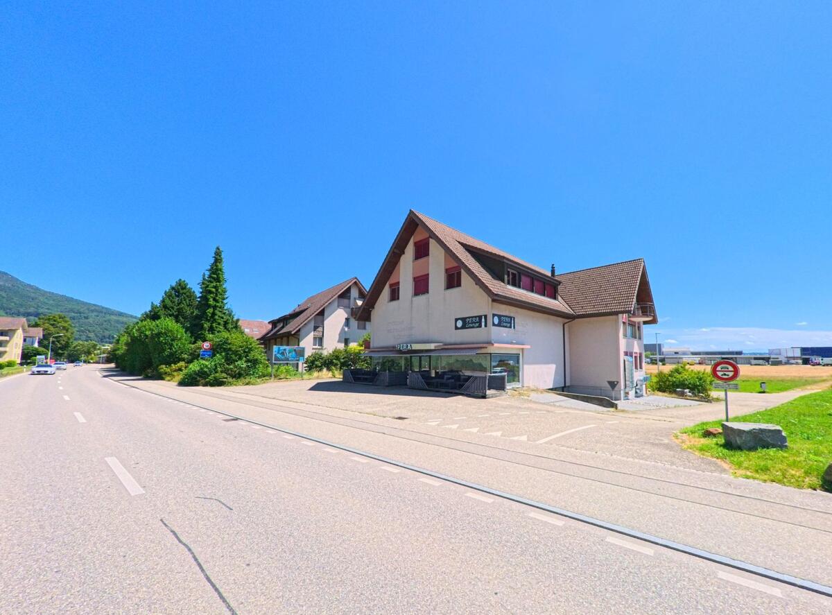 In Niederbipp (4704, Kanton Bern) steht ein Gebaeude mit steilem Satteldach an einer Ausfallstrasse, daneben beginnt ein Weg in Richtung offenes Feld.
