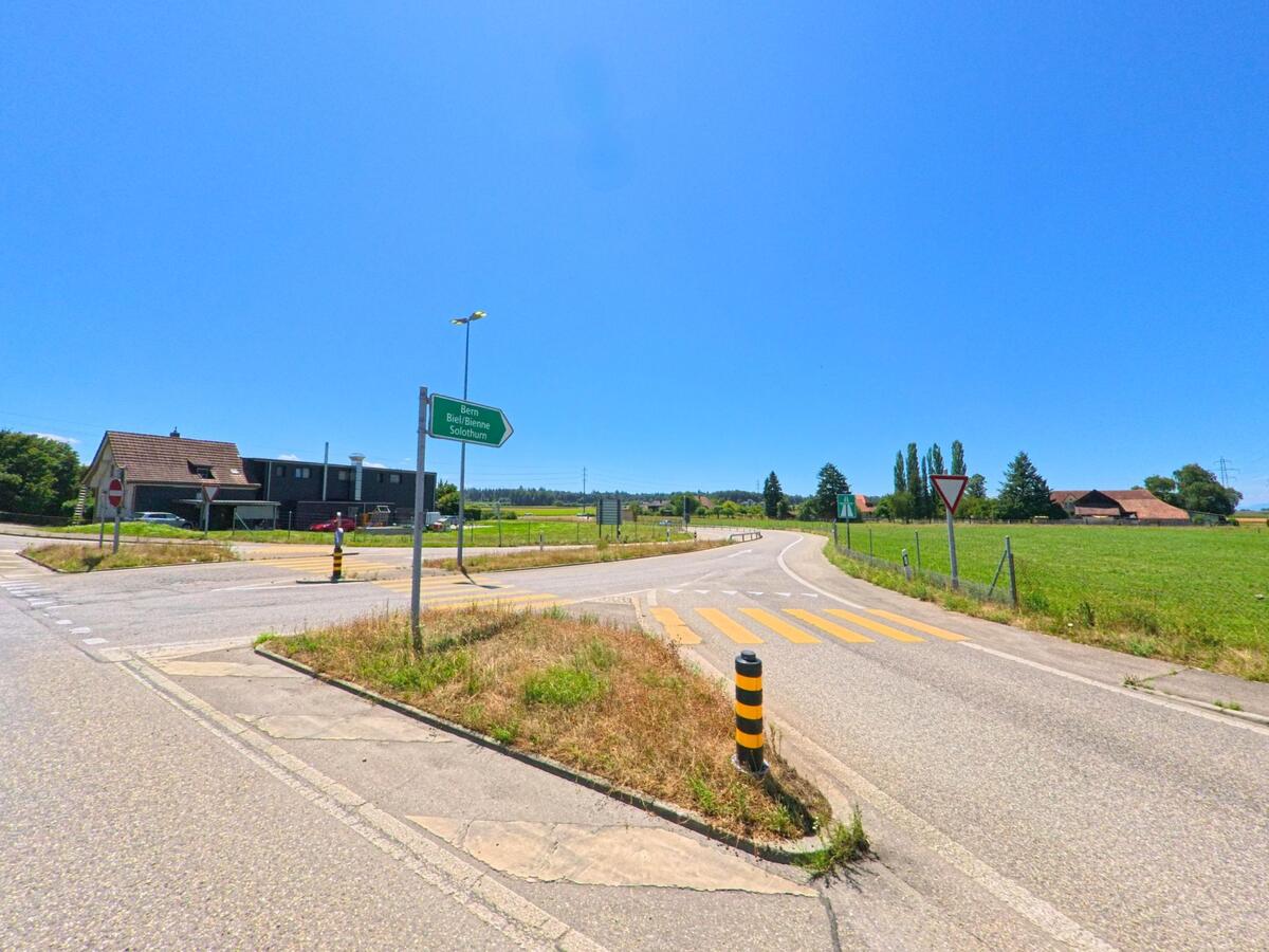 In Niederbipp (4704, Kanton Bern) fuehrt eine Landstrasse mit gelbem Fussgaengerstreifen vorbei an Wegweisern in Richtung Bern, Biel/Bienne und Solothurn durch offenes Gruenland.