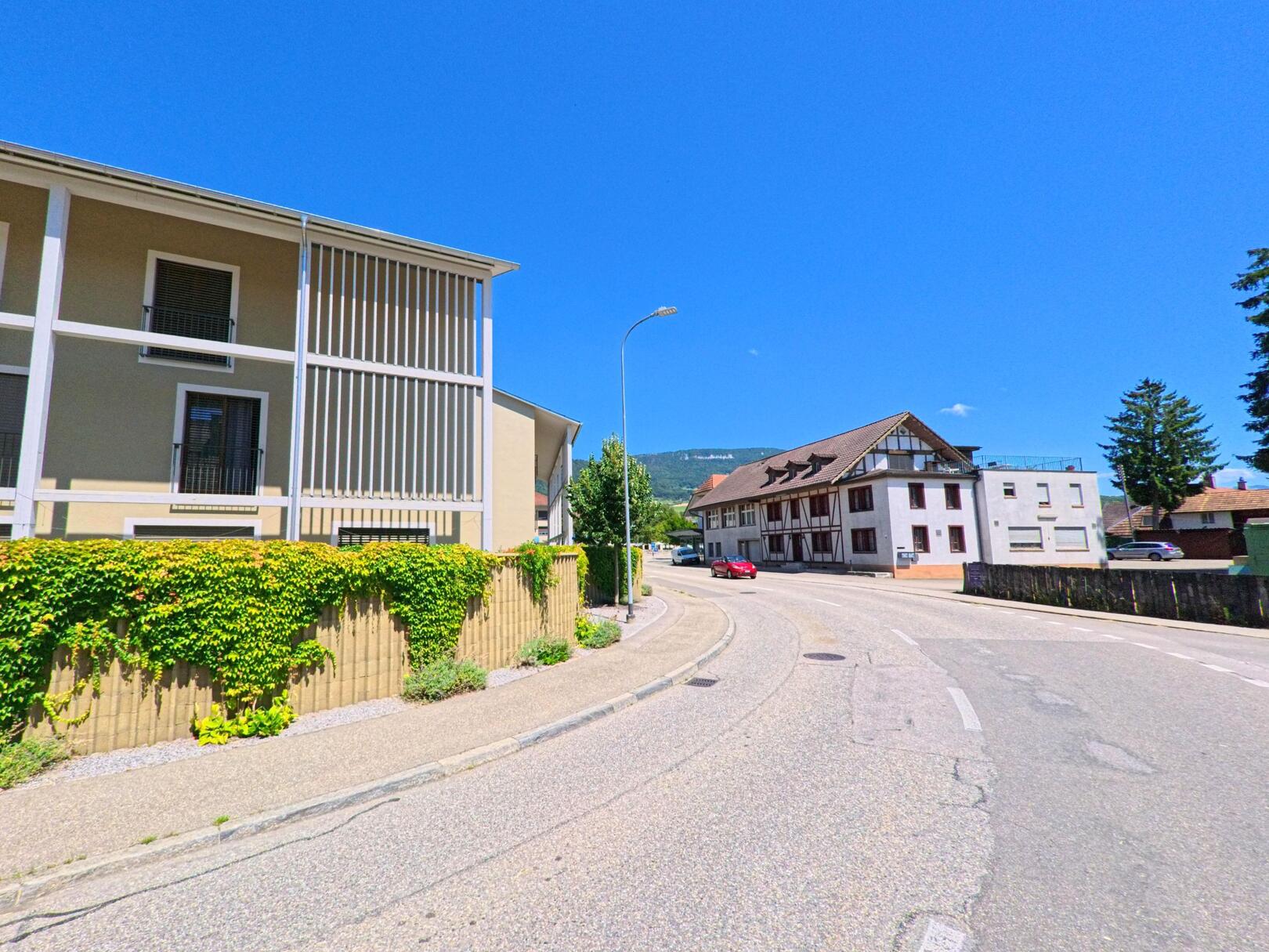 Eine sanft geschwungene Strasse in Niederbipp (4704, Kanton Bern) fuehrt an modernen und traditionellen Gebaeuden entlang unter klarem Sommerhimmel.