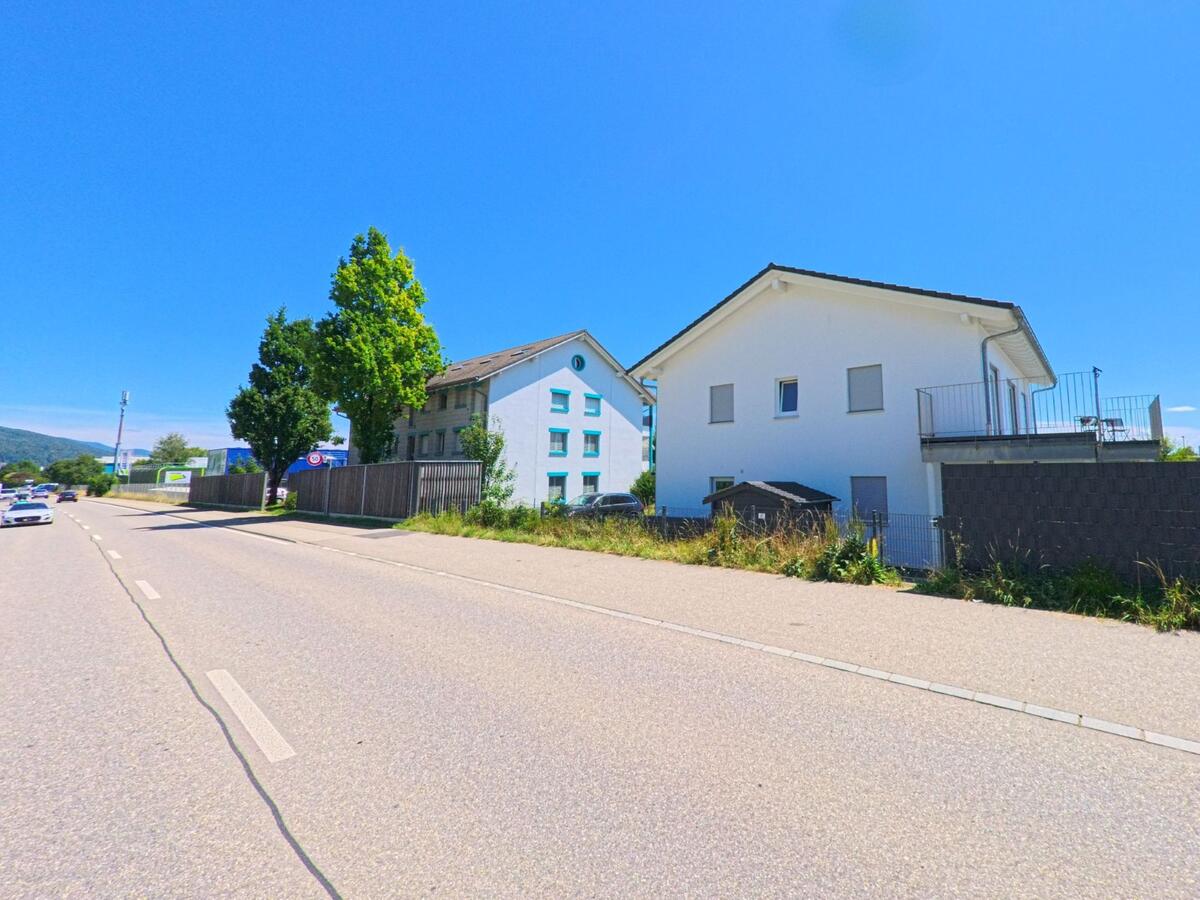 Entlang einer gut befahrenen Strasse in Niederbipp (4704, Kanton Bern) stehen moderne Wohnhaeuser mit gepflegtem Umfeld unter blauem Himmel.